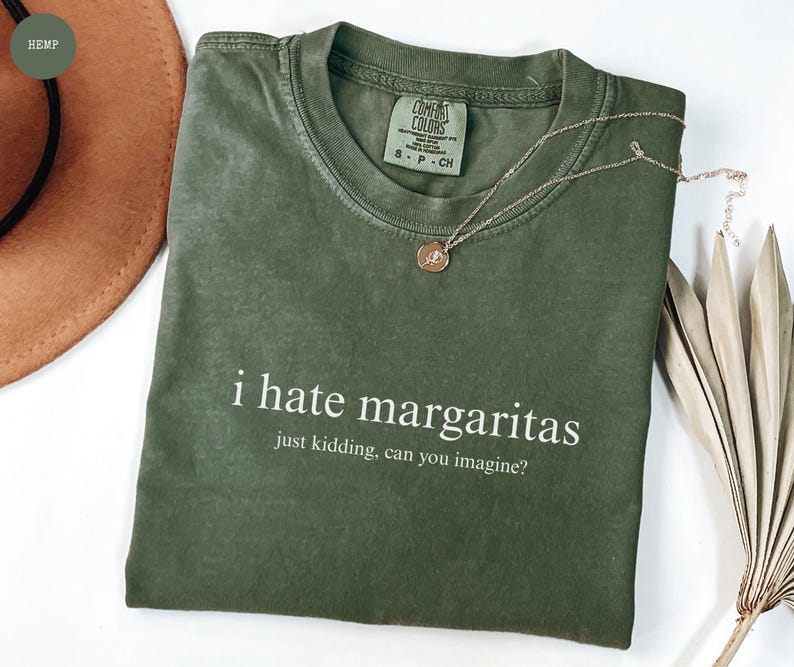 May include: A sage green short-sleeve t-shirt with a crew neckline. The shirt features white text that reads: i hate margaritas, just kidding, can you imagine? A thin gold-tone chain necklace with a small circular pendant rests on the collar.