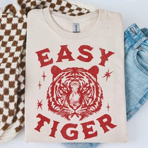 May include: Cream-colored t-shirt with the words "EASY TIGER" in red, along with a red tiger graphic. The shirt is folded, next to a brown and white checkered sweater and blue jeans.