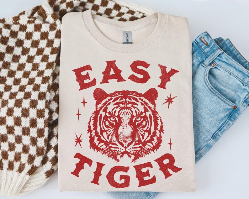 May include: Cream-colored t-shirt with the words "EASY TIGER" in red, along with a red tiger graphic. The shirt is folded, next to a brown and white checkered sweater and blue jeans.