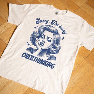 May include: Cream-colored t-shirt with a blue graphic of a woman and the text "Sorry, I'm busy overthinking." The design features a vintage illustration style with stars.