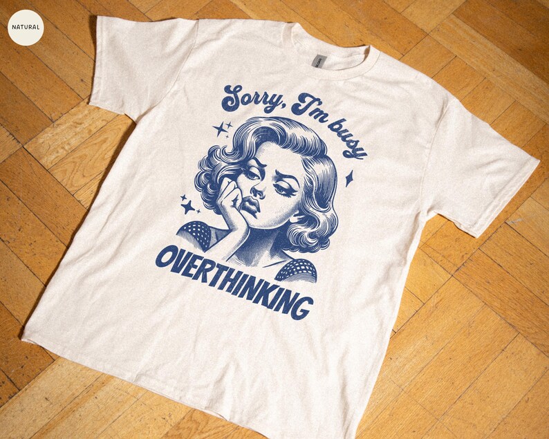 May include: Cream-colored t-shirt with a blue graphic of a woman and the text "Sorry, I'm busy overthinking." The design features a vintage illustration style with stars.