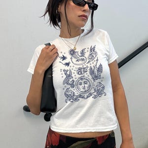 May include: White t-shirt with a navy blue illustration featuring celestial and mythical creatures. The design includes a sun with a face, dragons, and other figures. The shirt has short sleeves and a crew neck.