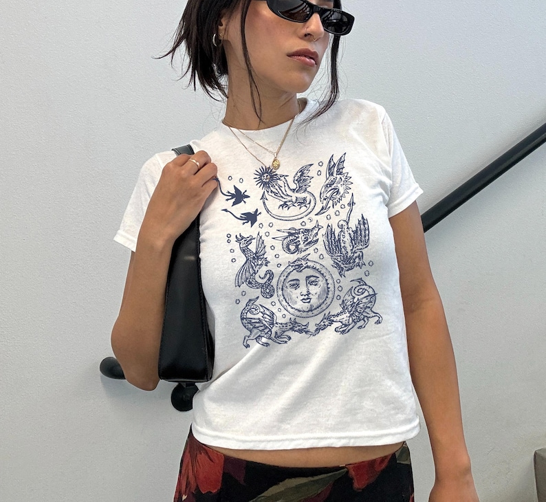 May include: White t-shirt with a navy blue illustration featuring celestial and mythical creatures. The design includes a sun with a face, dragons, and other figures. The shirt has short sleeves and a crew neck.