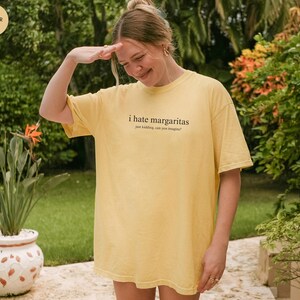 May include: A butter yellow short-sleeve t-shirt featuring the black text "i hate margaritas" followed by the smaller text "just kidding, can you imagine?" printed on the center of the chest. The garment has a crew neckline and a relaxed fit.