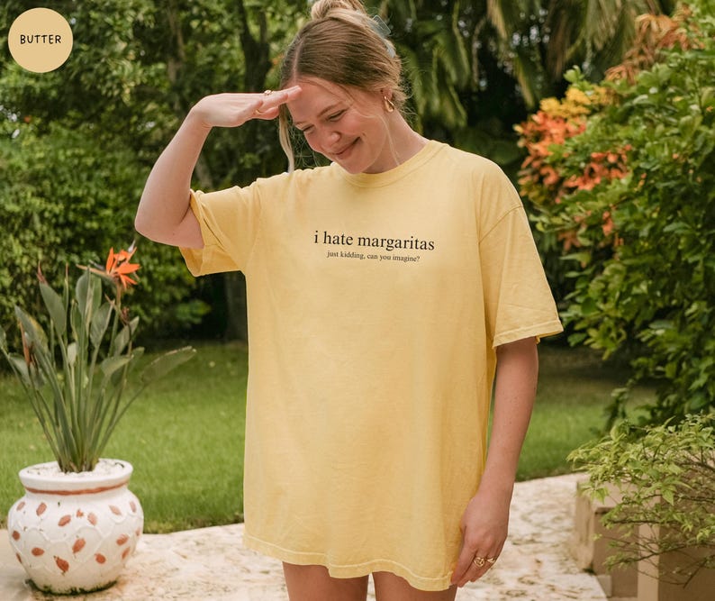 May include: A butter yellow short-sleeve t-shirt featuring the black text "i hate margaritas" followed by the smaller text "just kidding, can you imagine?" printed on the center of the chest. The garment has a crew neckline and a relaxed fit.