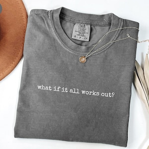 May include: A gray Comfort Colors t-shirt with the phrase "what if it all works out?" in white lettering. A silver necklace with a small pendant rests on the shirt. A brown hat and dried plant are in the background.