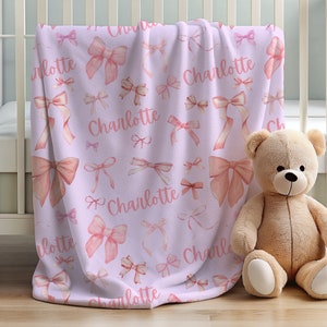 Pode incluir: Um cobertor de beb&ecirc; lil&aacute;s com um padr&atilde;o de la&ccedil;os rosa e o nome "Charlotte" em script rosa correspondente. Um urso de pel&uacute;cia bege est&aacute; sentado ao lado do cobertor, que est&aacute; sobre um ber&ccedil;o branco.