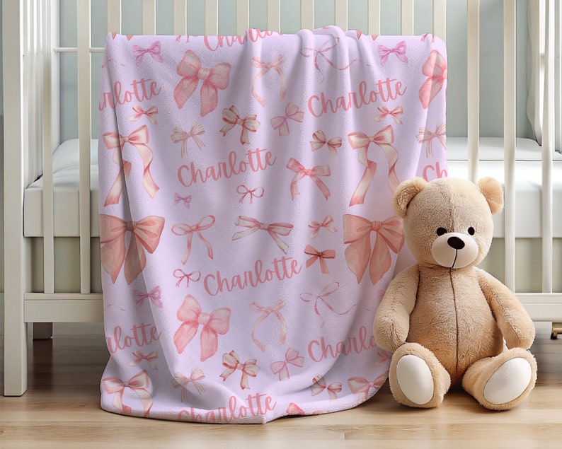 Pode incluir: Um cobertor de beb&ecirc; lil&aacute;s com um padr&atilde;o de la&ccedil;os rosa e o nome "Charlotte" em script rosa correspondente. Um urso de pel&uacute;cia bege est&aacute; sentado ao lado do cobertor, que est&aacute; sobre um ber&ccedil;o branco.