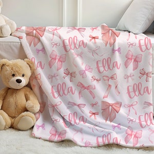 Pode incluir: Um cobertor branco com um padr&atilde;o repetido de la&ccedil;os rosa e o nome "Ella" em escrita rosa a condizer. Os la&ccedil;os variam em tamanho e estilo, criando um design suave e feminino. O cobertor &eacute; exibido numa cama.