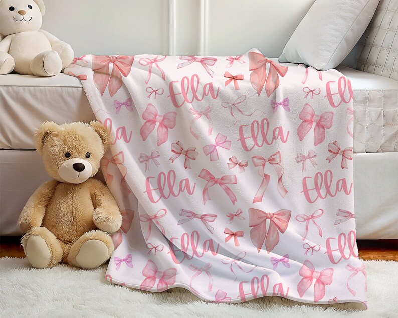 Pode incluir: Um cobertor branco com um padr&atilde;o repetido de la&ccedil;os rosa e o nome "Ella" em escrita rosa a condizer. Os la&ccedil;os variam em tamanho e estilo, criando um design suave e feminino. O cobertor &eacute; exibido numa cama.