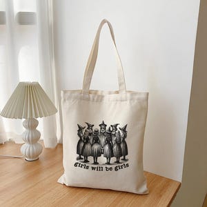 May include: A white canvas tote bag with a black and white illustration of a group of girls in witch costumes. The text "girls will be girls" is printed below the illustration.