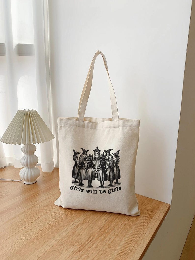 May include: A white canvas tote bag with a black and white illustration of a group of girls in witch costumes. The text "girls will be girls" is printed below the illustration.