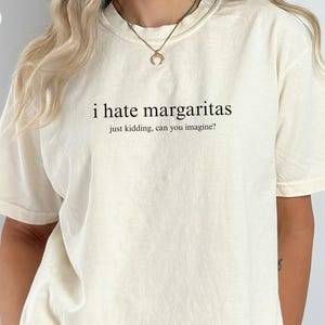May include: An ivory-colored short-sleeve t-shirt featuring the black printed text "i hate margaritas" followed by the smaller text "just kidding, can you imagine?" centered on the chest. The shirt has a crew neckline and a relaxed fit.