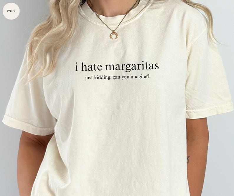 May include: An ivory-colored short-sleeve t-shirt featuring the black printed text "i hate margaritas" followed by the smaller text "just kidding, can you imagine?" centered on the chest. The shirt has a crew neckline and a relaxed fit.