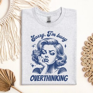 May include: Ash gray t-shirt with a blue graphic of a woman with the text "Sorry, I'm busy OVERTHINKING". The illustration features a vintage style with a woman resting her chin on her hand.