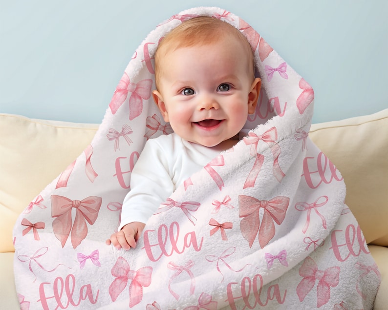 Pode incluir: Um beb&ecirc; envolto em um cobertor com capuz branco com um padr&atilde;o de la&ccedil;os rosa e o nome "Ella" em escrita rosa. O cobertor tem uma textura macia e felpuda e &eacute; drapeado sobre a cabe&ccedil;a do beb&ecirc;.