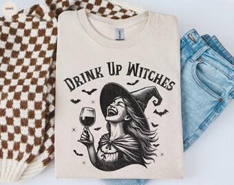 Puede incluir: Camiseta color arena con el texto "DRINK UP WITCHES" sobre una ilustración en blanco y negro de una bruja riendo que sostiene una copa de vino. El diseño incluye murciélagos y un sombrero de bruja. La camiseta está doblada junto a unos vaqueros azules y un suéter a cuadros marrón y blanco.