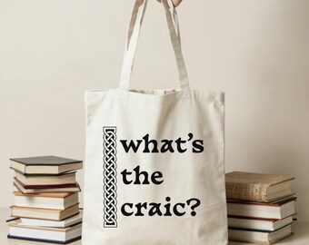 May include: Cream-colored tote bag with black text that reads "what's the craic?" and a black Celtic knot design. The bag is held by a hand and is next to a stack of books.