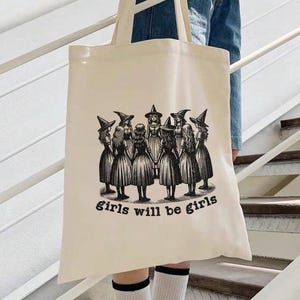 May include: A beige canvas tote bag with a black and white illustration of a group of witches holding hands in a circle. The text "girls will be girls" is printed below the illustration.
