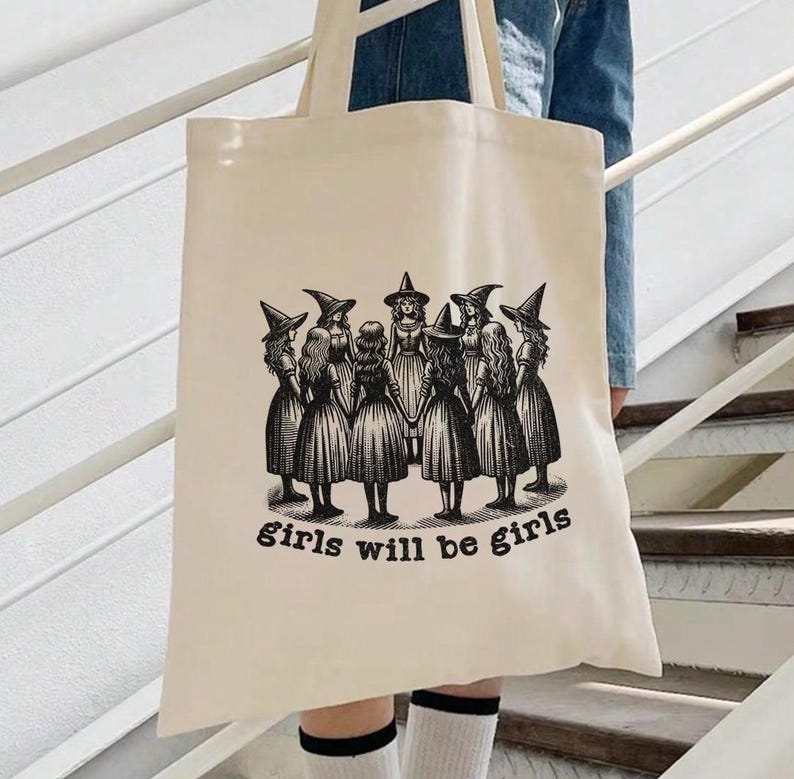 May include: A beige canvas tote bag with a black and white illustration of a group of witches holding hands in a circle. The text "girls will be girls" is printed below the illustration.