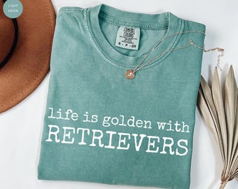 Puede incluir: Camiseta verde claro con el texto blanco "life is golden with RETRIEVERS". Un collar plateado con un colgante está sobre la camiseta. Un sombrero marrón y una planta seca están en el fondo.