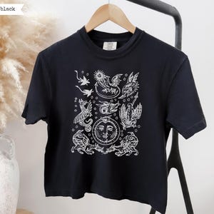 May include: A black short-sleeve t-shirt featuring a white graphic print of multiple dragons and a central sun-like face. The design includes various mythical creatures arranged in a cluster, with small circular accents surrounding the figures.