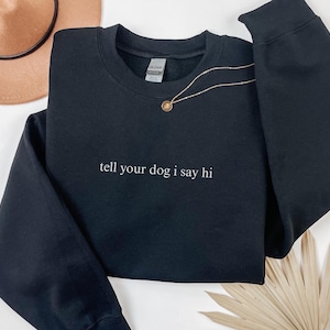 Puede incluir: Sudadera negra de cuello redondo con el texto blanco "tell your dog i say hi". Un collar dorado con un colgante está sobre la sudadera. Un sombrero marrón y una hoja de palma seca están en el fondo.