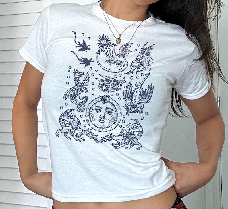 May include: White t-shirt featuring a dark blue illustration of mythical creatures and celestial bodies. The design includes dragons, a sun, a moon with a face, and other fantastical figures. The shirt has short sleeves and a crew neck.