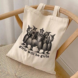 May include: A white canvas tote bag with black handles and a black and white illustration of a group of witches with the text "girls will be girls".