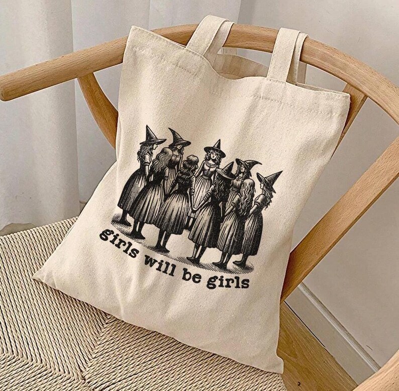 May include: A white canvas tote bag with black handles and a black and white illustration of a group of witches with the text "girls will be girls".