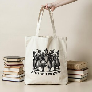 May include: A white canvas tote bag with a black and white illustration of a group of witches in a circle. The text "girls will be girls" is printed below the illustration.