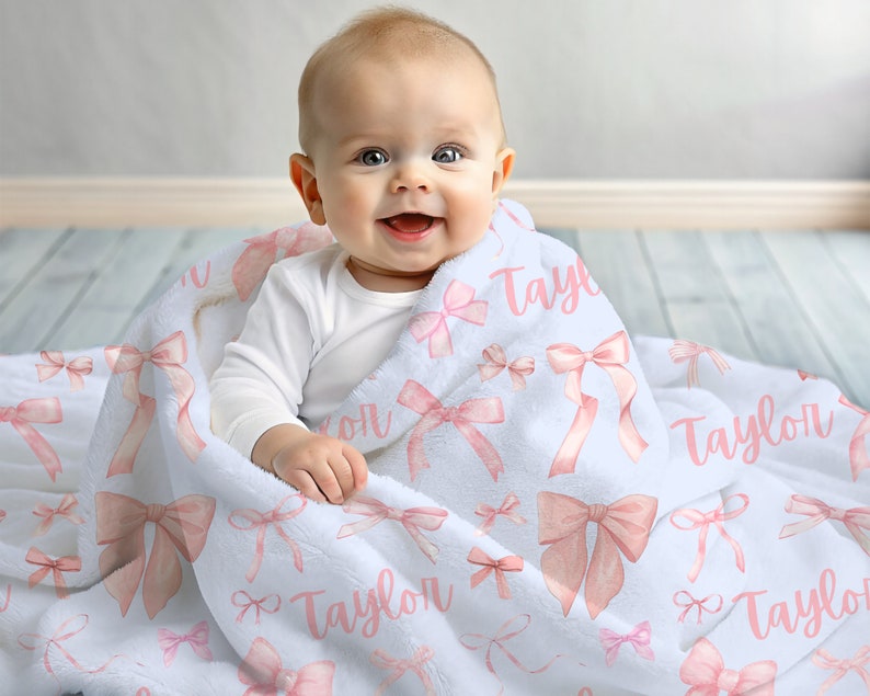 Pode incluir: Um cobertor branco para beb&eacute; com um padr&atilde;o de la&ccedil;os cor-de-rosa e o nome "Taylor" em escrita rosa. O cobertor est&aacute; enrolado em volta de um beb&eacute; com um body branco.