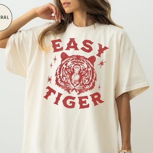 May include: Cream-colored t-shirt with a red tiger graphic and the words "EASY TIGER". The design includes a tiger's face and starburst accents. The shirt has short sleeves and a relaxed fit.