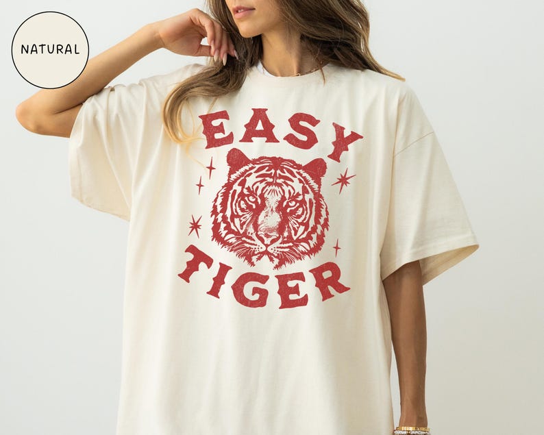 May include: Cream-colored t-shirt with a red tiger graphic and the words "EASY TIGER". The design includes a tiger's face and starburst accents. The shirt has short sleeves and a relaxed fit.