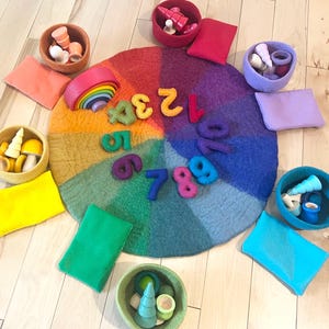 May include: A circular rainbow-colored felt mat featuring felt numbers one through ten arranged in a circle. Surrounding the mat are six felt bowls in various colors, each containing small wooden figures, along with six rectangular felt pouches.