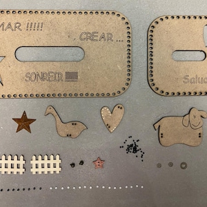 May include: A set of wooden craft supplies for making a bag or purse. The set includes two large pieces with cutouts for handles and text that reads "AMAR!!!", "CREAR...", "SONREIR", and "Salud!!!". The set also includes smaller pieces like a heart, a star, a fence, a goose, and a sheep.