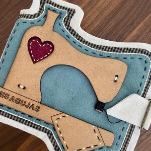 Puede incluir: Un alfiletero de fieltro azul con forma de máquina de coser con base de madera marrón. El alfiletero tiene un corazón rojo en la parte superior y las palabras "Mis Agujas" en el lateral.