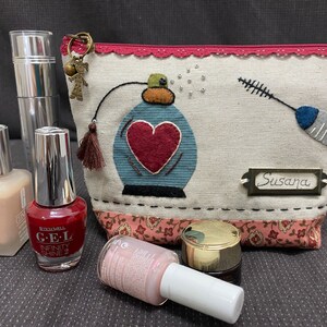 Può includere: Una trousse da toilette in tessuto con motivo toile rosso, bianco e blu con un design a forma di cuore e il nome "Susana" su un'etichetta metallica. La trousse è piena di vari prodotti di bellezza, tra cui un flacone di smalto rosso, un flacone di smalto trasparente, un flacone di fondotinta e un piccolo barattolo di crema.