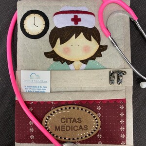 Op de afbeelding: Een beige stoffen boek met een rood kruis en een cartoonverpleegster op de voorkant. Het boek heeft een roze stethoscoop eromheen gewikkeld en een bruin ovaal label met de tekst "CITAS MEDICAS".