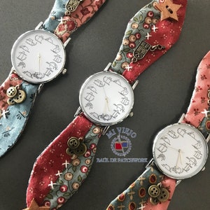 Op de afbeelding: Drie zilveren polshorloges met witte wijzerplaten en zwarte wijzers. Elk horloge heeft een andere gepatroneerde stoffen band met verschillende versieringen, waaronder knopen, sterren en bedels. De banden zijn in tinten roze, rood en blauw.