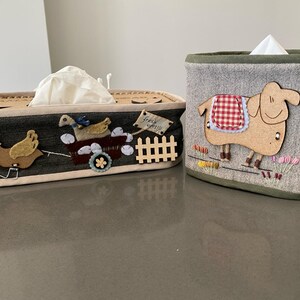 Puede incluir: Dos cubiertas de caja de pañuelos de tela con detalles bordados. Una presenta una oveja marrón y blanca con una silla de montar de cuadros rojos y blancos. La otra presenta un ganso marrón y blanco tirando de un carro con un ganso marrón y blanco y un pollito marrón y blanco dentro. Ambas cubiertas tienen un fondo gris con un ribete blanco.