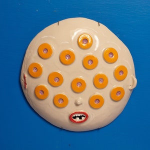 May include: A white ceramic sculpture of a face with multiple orange eyes and a red mouth. The sculpture is against a blue background.