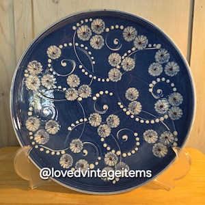 May include: A round ceramic plate with a deep blue glaze featuring a hand-painted pattern of white daisy-like flowers, swirling lines, and small white dots. The text @lovedvintageitems is printed in white at the bottom of the plate.