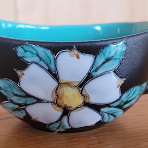 Puede incluir: Cremera de cerámica negra con interior y asa turquesa. El exterior está decorado con un diseño floral blanco con detalles amarillos y verdes. La cremera está sobre una superficie de madera.