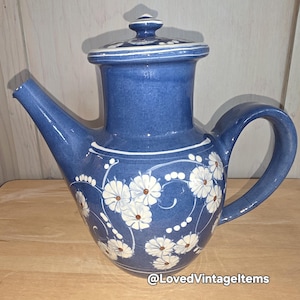 Peut inclure: Une théière en céramique bleue avec un couvercle assorti, ornée d'un motif floral blanc peint à la main avec de petits centres bruns et des vignes pointillées blanches. La théière a une anse courbée, un bec verseur et le texte @LovedVintageItems.