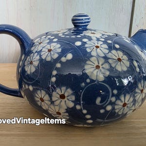 May include: A round ceramic teapot with a deep blue glaze featuring a white daisy floral pattern and brown centers. The teapot has a matching lid with a knob, a curved handle, and a spout. The text @LovedVintageItems is visible at the bottom.