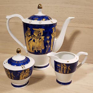May include: A white porcelain tea set with gold and blue accents. The set includes a teapot, creamer, and sugar bowl. The design features Egyptian-inspired figures and hieroglyphics.