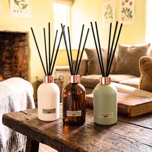 May include: Three reed diffusers with black reeds and copper-coloured collars are displayed on a rustic wooden table. The diffusers are in white, amber, and sage green bottles, each labelled "SCENT GROVE". The background includes a fireplace and a sofa.