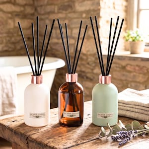 May include: Three reed diffusers on a wooden surface. The diffusers are in white, amber, and green glass bottles with copper-coloured collars and black reeds. Each bottle has a label that reads "SCENT GROVE".