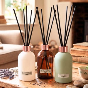 May include: Three reed diffusers in white, brown, and green glass bottles with copper-coloured collars and black reeds. Each bottle has a label that reads "SCENT GROVE". The diffusers are arranged on a wooden surface.